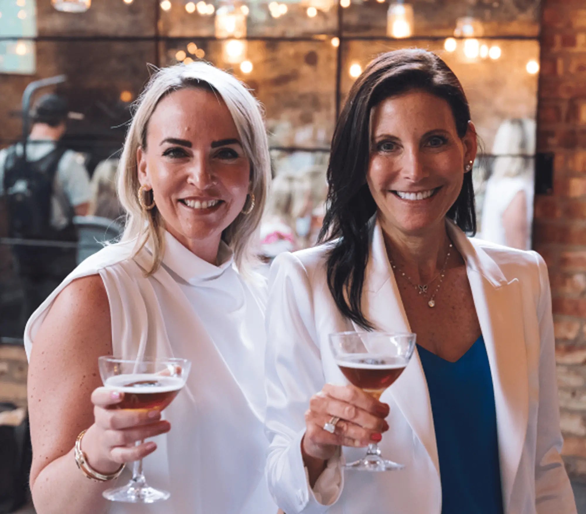 Mara Smith and Erika Thieme (National Sales & Marketing Director at Inspiro Tequila) holding drinks in a social setting with brick wall and blurred people in the background