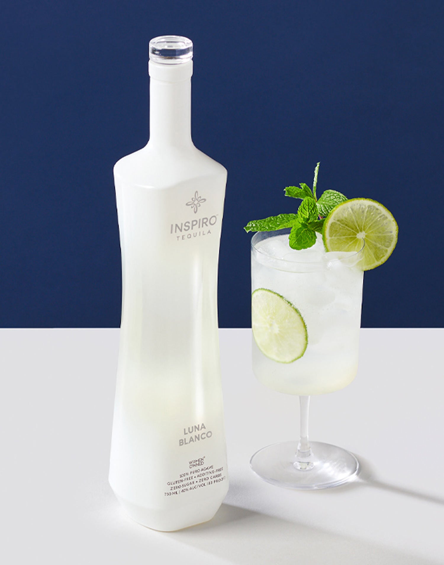 Bottle of Inspiro Tequila with a cocktail in a glass garnished with lime and mint on a white surface.