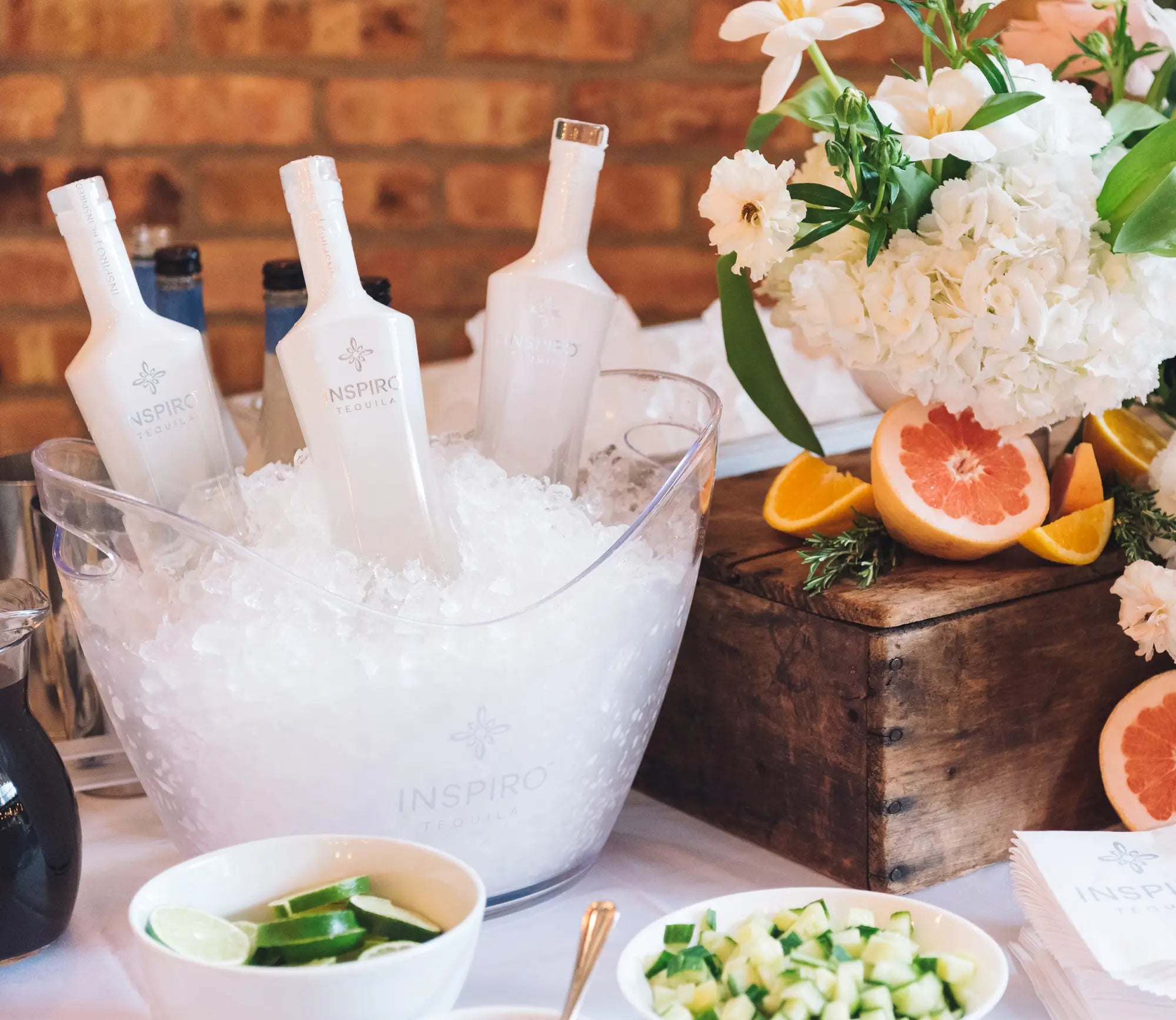 Bottles of Inspiro Tequila in an ice bucket with a floral arrangement and fruit on a table.