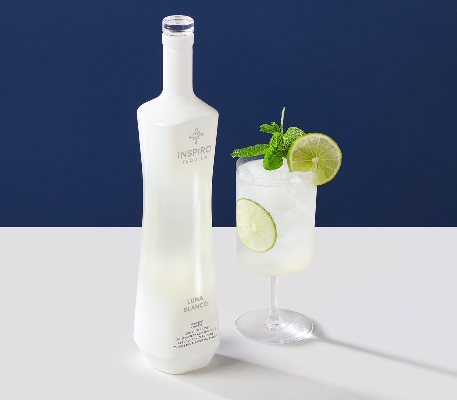 Bottle of Inspiro Luna Blanco with a cocktail in a glass garnished with lime and mint on a white surface with a dark blue background.