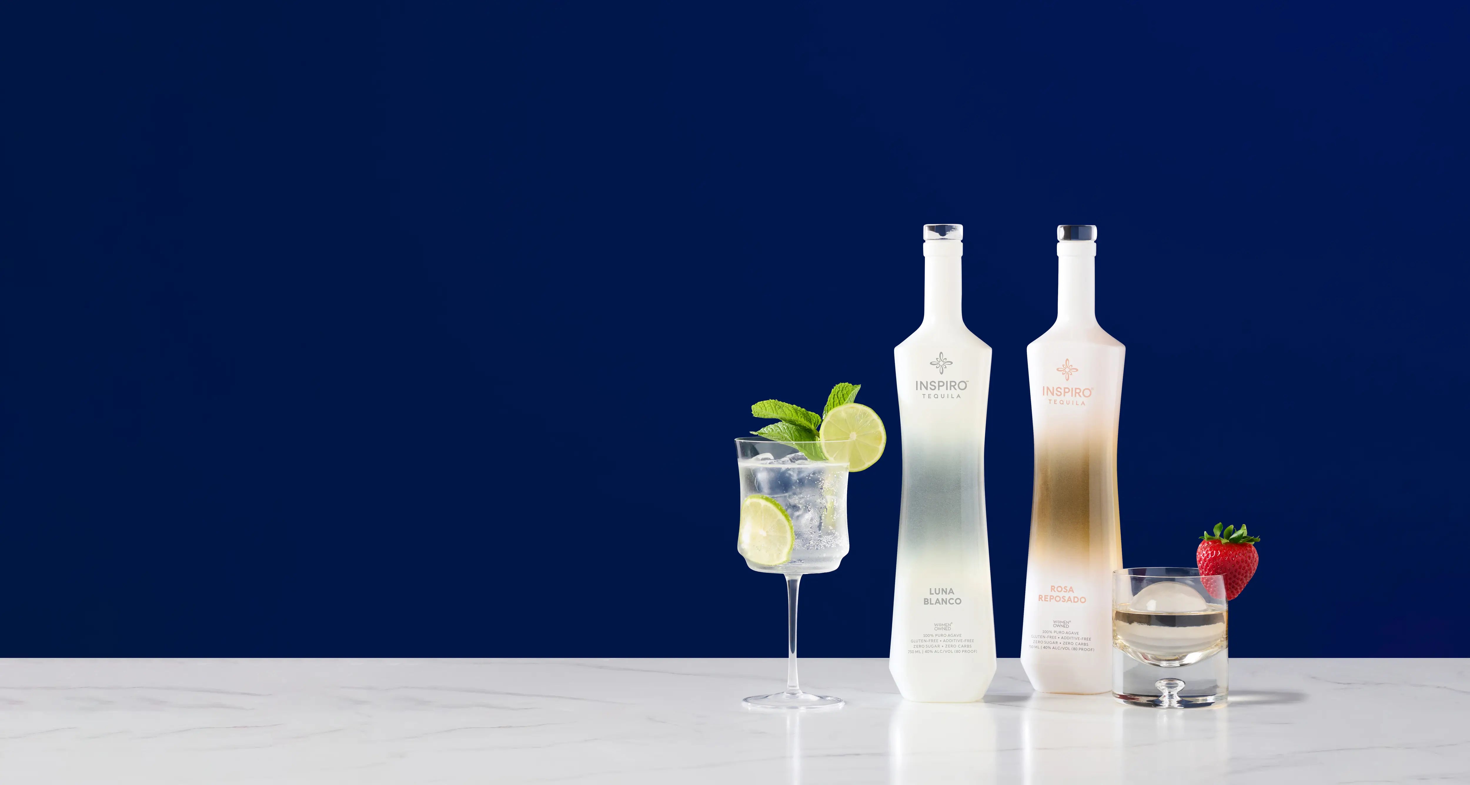 Two bottles of Inspiro Tequila with a glass of water and garnishes on a white surface against a dark blue background