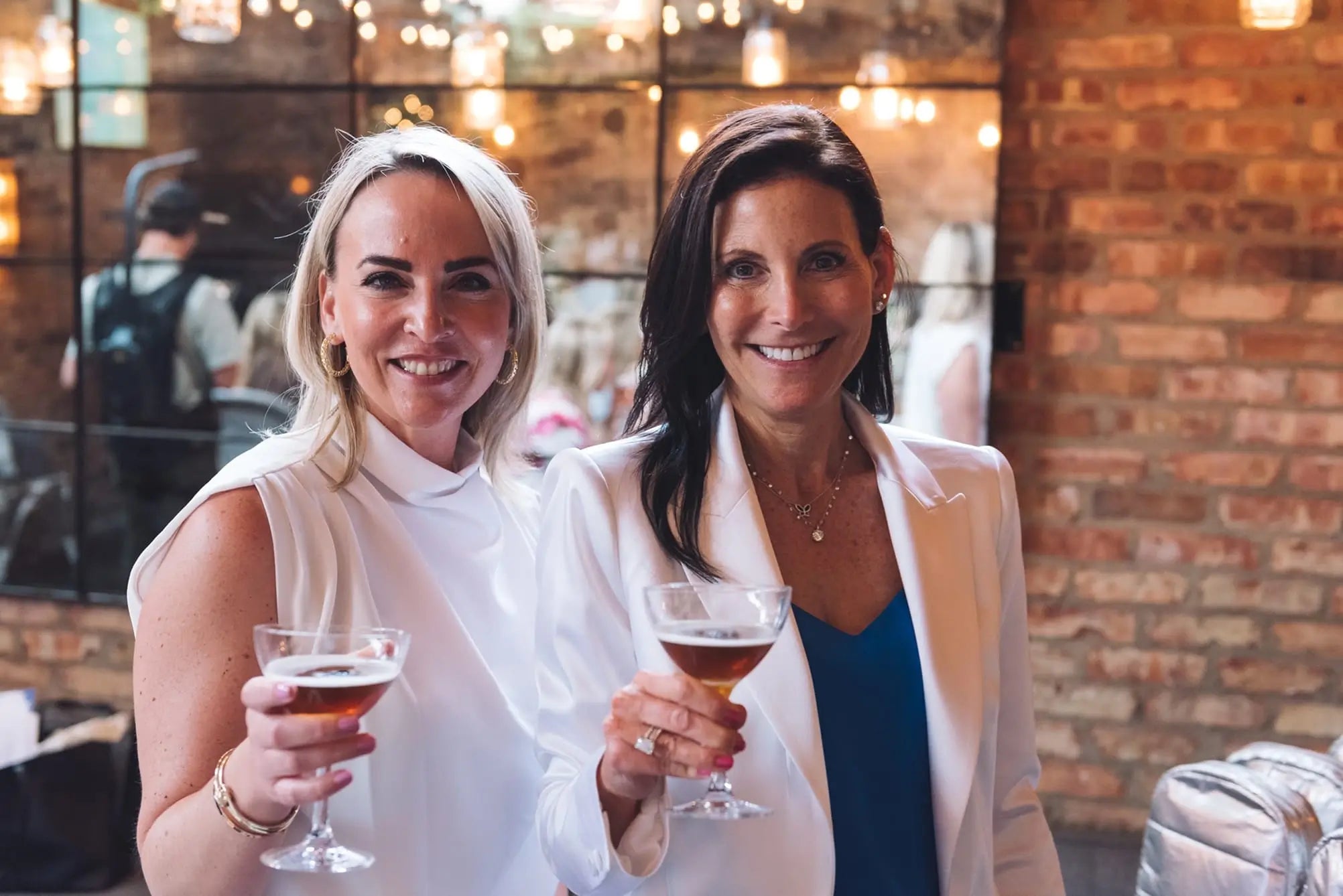 Mara Smith and Erika Thieme (National Sales & Marketing Director at Inspiro Tequila) holding drinks in a social setting with brick wall and blurred people in the background