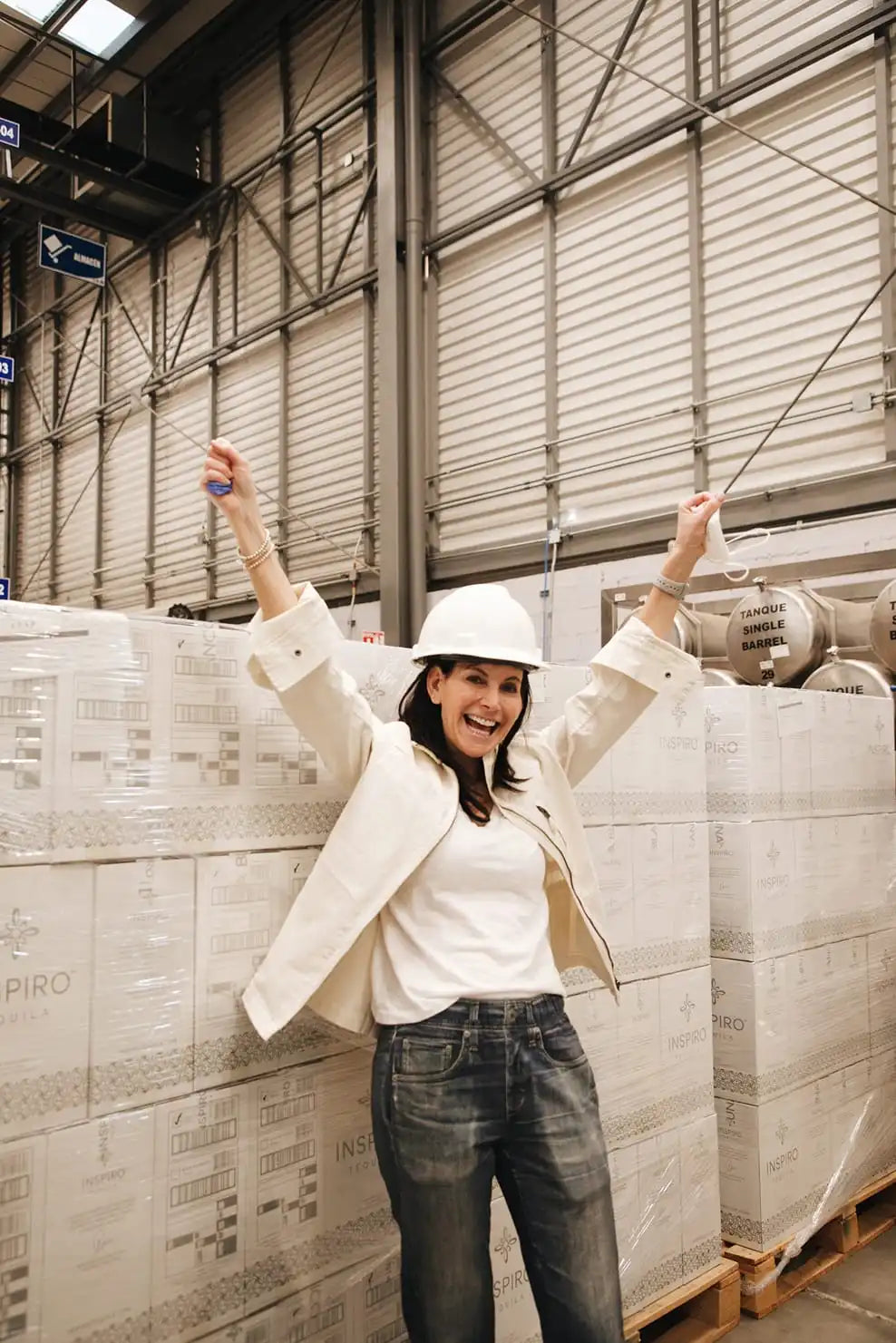 Mara Smith in a warehouse wearing a hard hat and white jacket, surrounded by stacked boxes of Inspiro Tequila