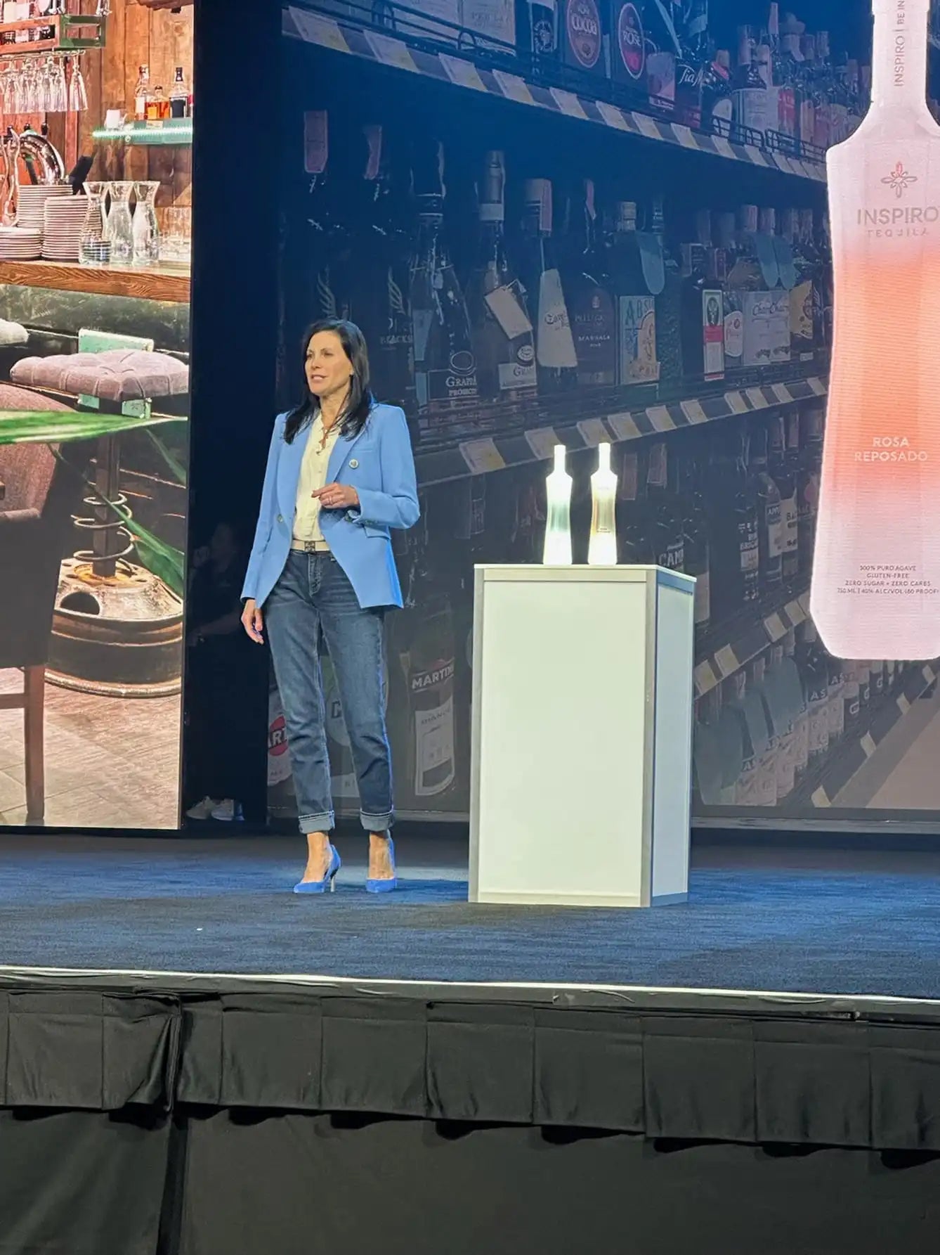 Mara Smith in blue blazer standing on a stage with a backdrop of Inspiro Tequila bottles and a podium.